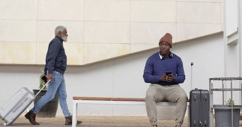 African American travelers waiting on bench with luggage and headphones in urban station
