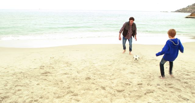 Father and son are spending quality time together on the beach, playing soccer by the ocean. The waves are gently rolling in, and both are barefoot, fully engaged in the activity. Great for themes of family bonding, beach vacations, outdoor activities, and child-parent relationships.