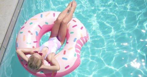 Young girl relaxing on pink donut float in backyard turquoise pool on sunny day