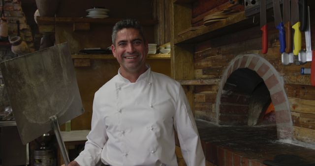 Smiling chef holding a large pizza peel in a rustic-style pizzeria with a brick oven. This warm and inviting setting highlights the traditional aspects of preparing Italian cuisine. Ideal for use in articles, blogs, and websites related to cooking, restaurants, and culinary professions.