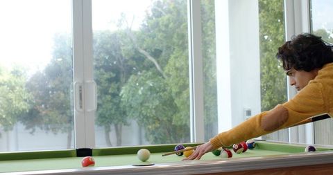 Young man leaning over pool table aiming cue ball in sunlit home game room