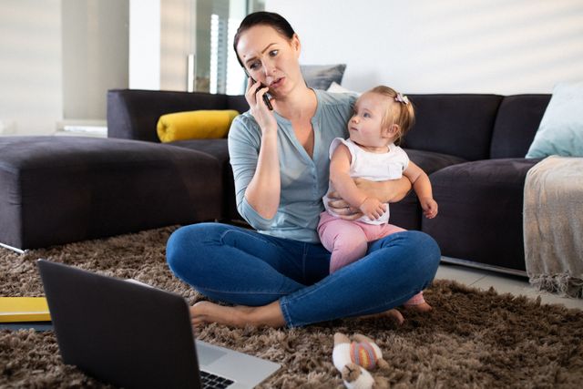 Caucasian mother sitting on floor holding baby daughter while talking on smartphone and using laptop. Ideal for articles on remote work, parenting during quarantine, balancing work and family life, and the challenges of working from home.