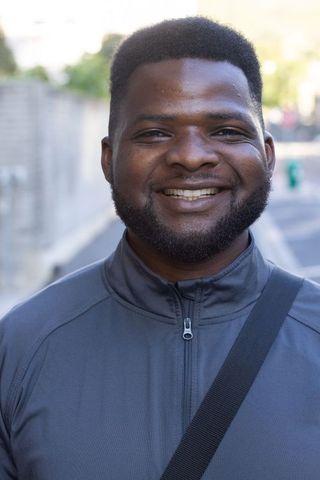African man smiling on urban sidewalk wearing gray zip-up jacket with crossbody strap