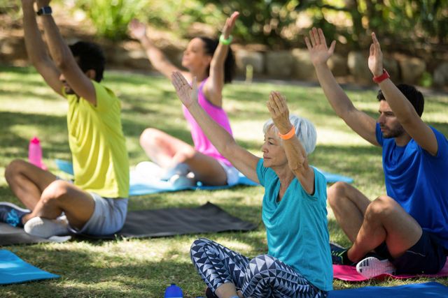Group of people practicing yoga in a park on a sunny day. They are sitting on yoga mats with arms raised, focusing on their practice. This image can be used for promoting outdoor fitness activities, wellness programs, yoga classes, and healthy lifestyle campaigns.
