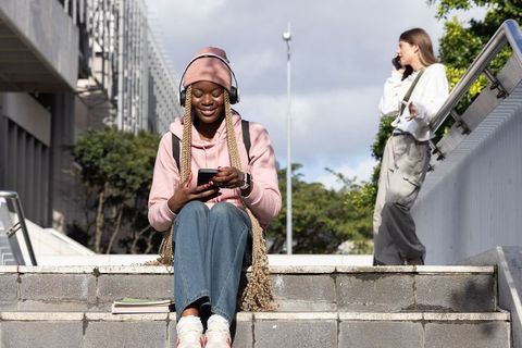 Black student in pink hoodie sitting on campus steps using phone and headphones with white peer on c