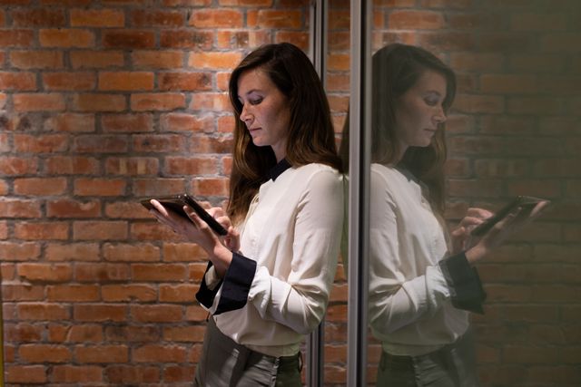Side view of a Caucasian businesswoman working in a modern creative office, wearing a white shirt using a digital tablet, leaning against a glass door, brick wall in the background.