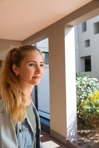 Young woman standing in sunlit covered walkway gazing thoughtfully wearing denim jacket