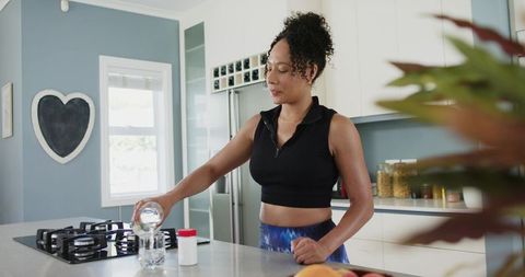 African american woman pouring water into glass on modern kitchen island for wellness