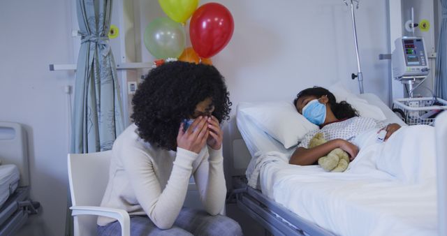 A woman sits beside a girl resting in a hospital bed with medical equipment and balloons in the background. The woman is showing concern while both are wearing masks. This image is ideal for use in articles or brochures related to healthcare, emotional support, pediatric care, hospital visits, and recovery processes. It can also be used in campaigns promoting the importance of family support during illness and hospital stays.