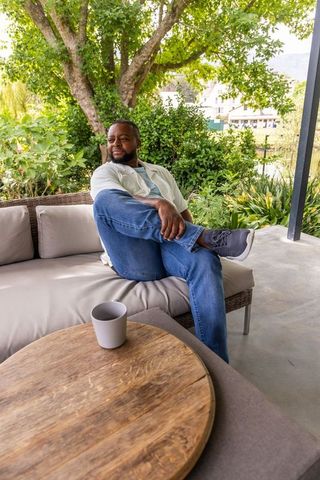 Relaxing african man sitting cross-legged on wicker sofa on sunlit leafy terrace with coffee mug