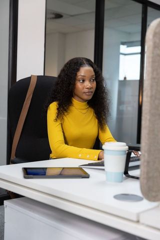 Focused woman typing at modern office desk with tablet, travel cup and high-back chair