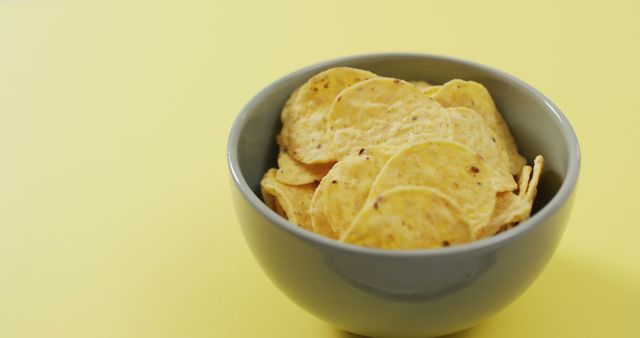 Stack of crispy potato chips in a grey bowl on a bright yellow background, showcasing a simple snack concept. Ideal for use in advertisements, food blogs, or menus highlighting simple and delicious snack options.
