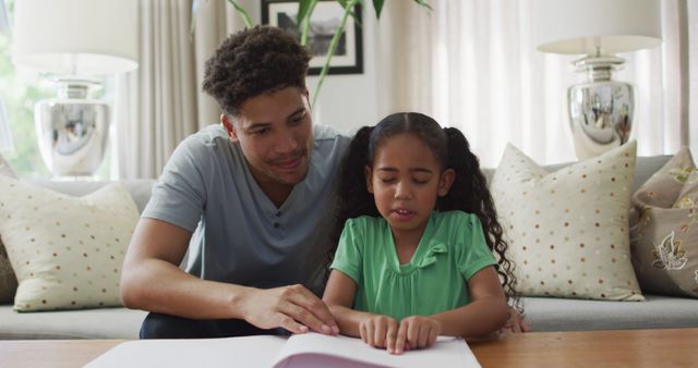 A caring father is helping his distressed child with homework at home. The girl seems frustrated while her father offers support and patience. This scene can be used in parenting blogs, educational materials, family support resources, and advertisements emphasizing parental involvement in children's education and emotional support.