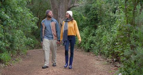 Couple walking hand-in-hand on leafy woodland trail wearing knit hats and rain boots