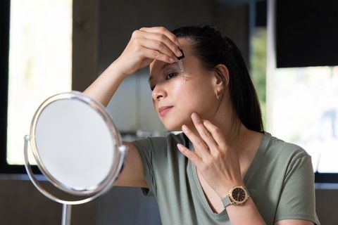 Asian woman applying facial serum with dropper at vanity mirror, natural skincare routine
