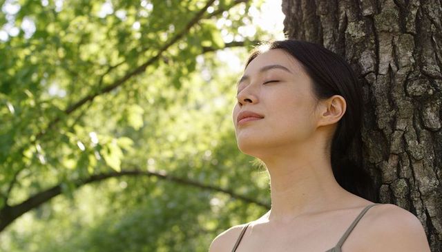 Asian woman leaning against tree with closed eyes enjoying peaceful sunlight and nature