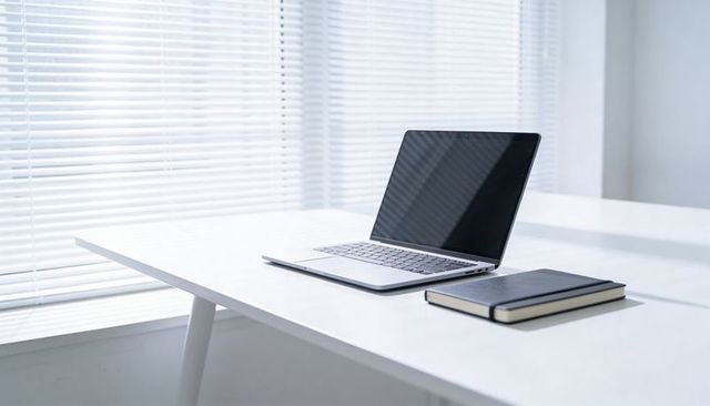Minimalist workspace with silver laptop and black notebook on white desk by window blinds
