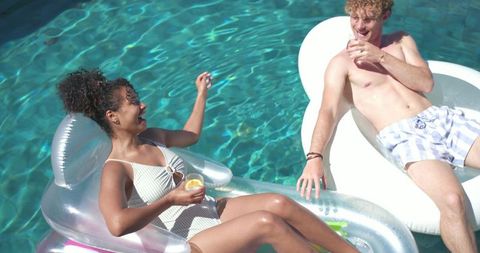 Young couple relaxing on inflatable floats in sunny backyard pool holding drinks