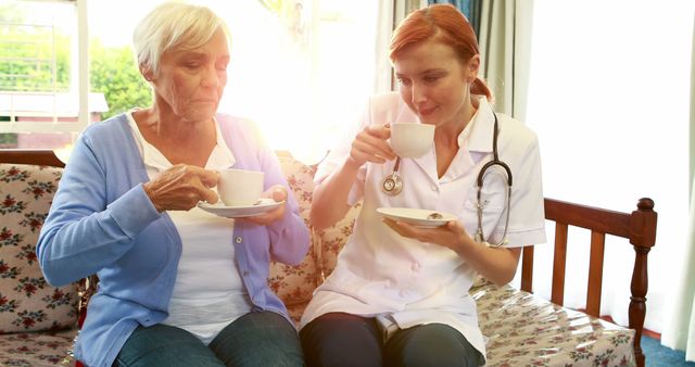 Nurse is spending quality time with an elderly woman, both enjoying a cup of tea on a sofa. This image depicts care and companionship provided by healthcare professionals to seniors in home settings. Suitable for illustrating home healthcare services, geriatric care, and medical staff proficiency in personal patient care.