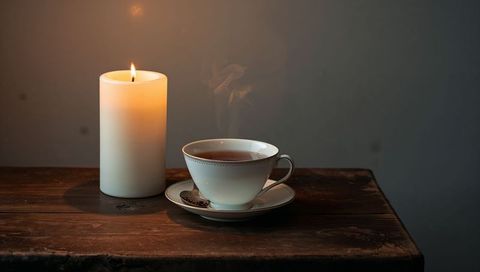 Glowing pillar candle and steaming porcelain teacup on rustic wooden table