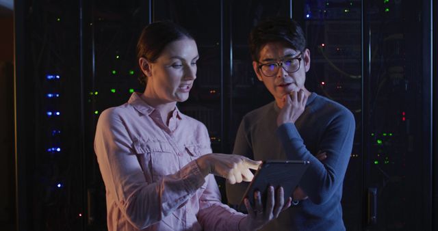 Two IT professionals are collaborating and discussing data displayed on a digital tablet in a server room. The scene is dimly lit by the lights of the servers, indicating a high-tech and focused environment. This visual can be used for content related to information technology, data analysis, teamwork in tech environments, and server management.