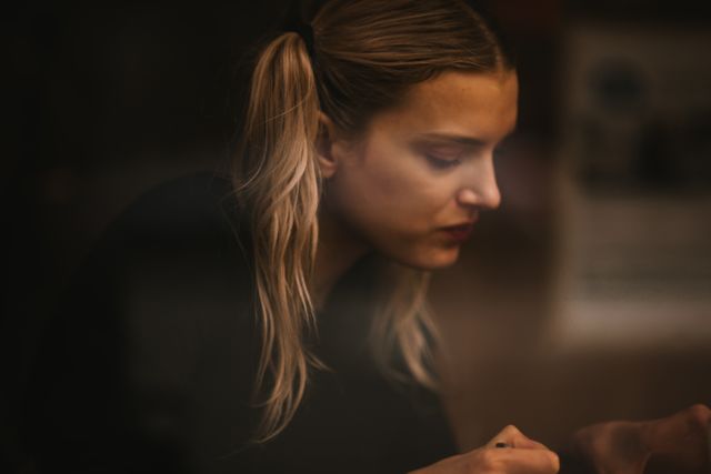 This photo captures a side profile of a pensive woman with blond hair in a dimly lit room. She appears deep in thought, offering a serene and moody feel. This image can be used for articles on introspection, mental health awareness, or any content that conveys contemplation and tranquility.