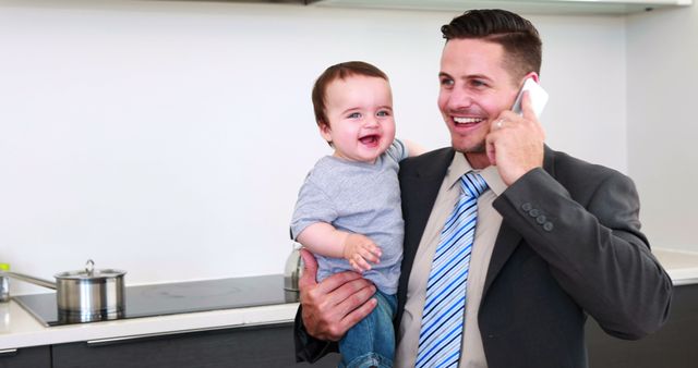 Father holding his baby son before work and talking on phone at home in the kitchen