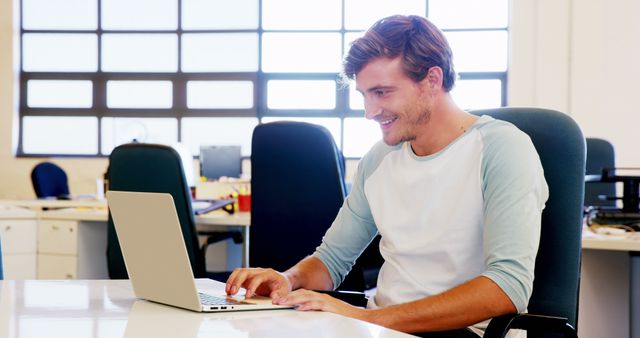 Young male professional focusing on laptop in a modern office while smiling. Ideal for use in articles or advertisements about workplace productivity, remote working, technology use in business, or office environment themes.