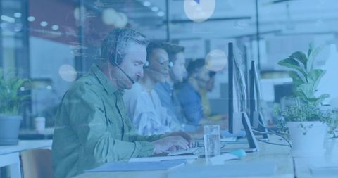 Mature support agent typing with headset in open-plan office, teamwork and productivity