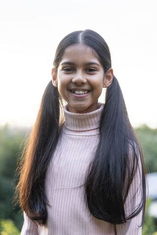 Smiling indian preteen girl wearing pink ribbed turtleneck, long pigtails, outdoor backlighting