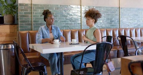 Black woman and mixed-race friend chatting over coffee in modern cozy urban cafe banquette