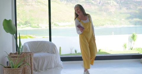 Mature woman dancing with smartphone in yellow overalls by sunlit ocean-view window