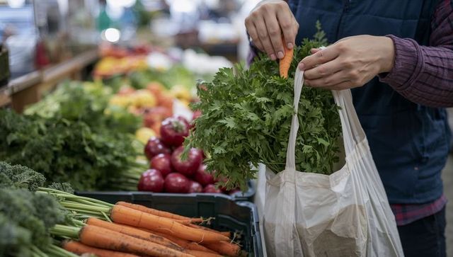 Shopper selecting carrot placing into reusable bag with leafy greens at farmers market