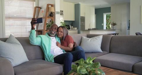 Couple taking selfie on gray sectional sofa in modern cozy living room with open kitchen
