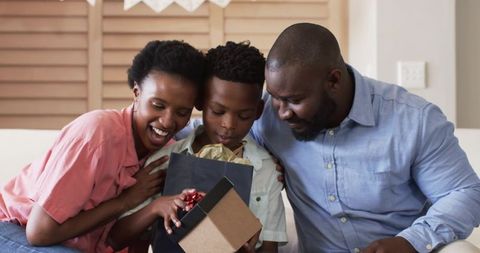 African American family celebrating son opening present on cozy living room sofa