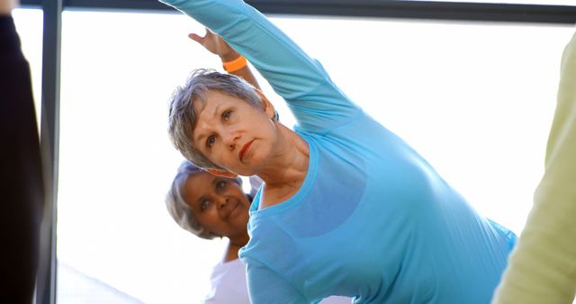 A middle-aged Caucasian woman in a blue top is stretching her arm during a yoga class, with copy space. Behind her, an African American woman follows the routine, highlighting the diversity and focus on wellness in the group.