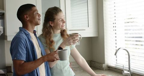 Sunlit mixed-race couple enjoying coffee by kitchen window during cozy morning