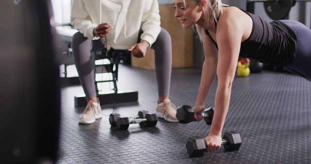 Image of diverse female fitness trainer encouraging woman raising dumbbells working out at a gym. Exercise, fitness and healthy lifestyle.