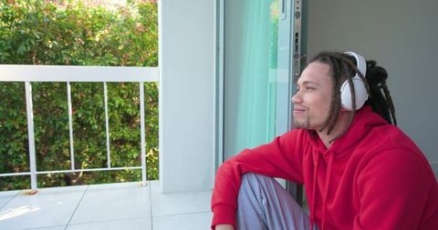 African american man listening to music on balcony doorway wearing red hoodie headphones
