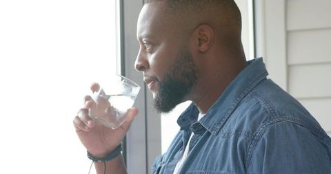 African american man drinking water by window, contemplative sunlit moment in denim