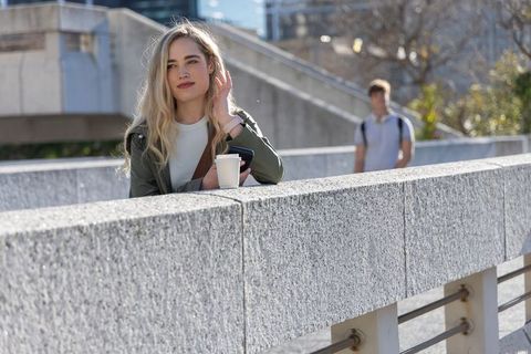 Young woman leaning on city overpass railing holding coffee and phone while commuter walks