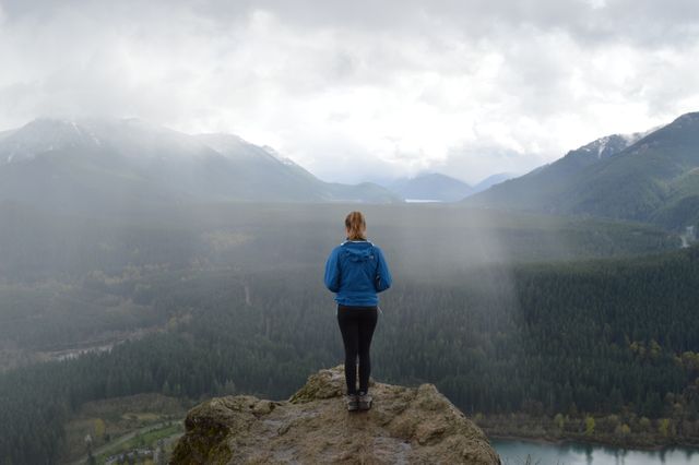 Person standing on a mountain cliff, overlooking a scenic valley covered in forests and mountains. Ideal for themes related to adventure, nature, exploring the outdoors, freedom, and tranquility. Suitable for travel blogs, outdoor sports advertisements, tourism brochures, and motivational content.