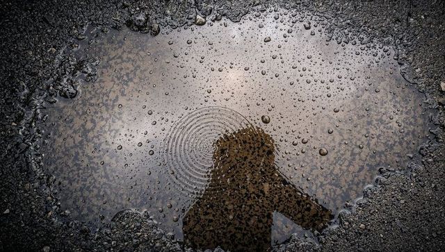 Hooded figure reflecting in rain puddle with ripples on pebbled asphalt