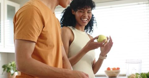 Laughing diverse couple preparing smoothie together in sunlit kitchen with fresh fruit