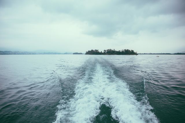 Boat wake captured on vast, calm ocean with an island in distance under an overcast sky. Ideal for themes related to tranquility, travel, and nature. Suitable for travel brochures, nature documentaries, and promotional materials on serene journeys or maritime experiences.