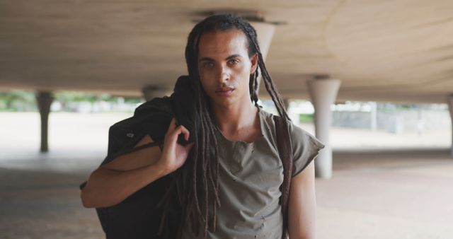 Young man with long dreadlocks carrying jacket over shoulder in contemporary urban area. He is wearing casual clothing, giving a confident and stylish look. Use for fashion articles, urban lifestyle blogs, or youth culture promotions.
