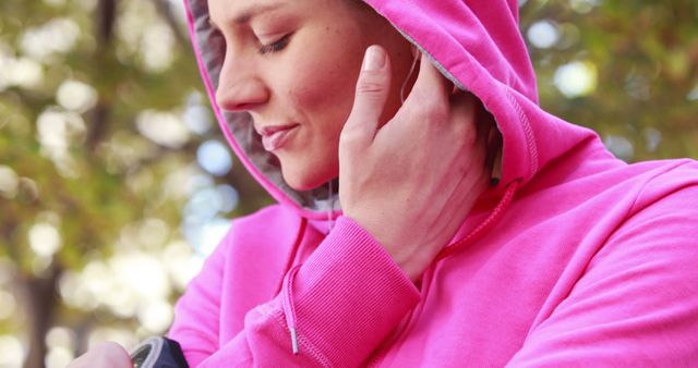 A young Caucasian woman in a pink hoodie is focused on her smartphone, with copy space. She appears to be engaged in a conversation or listening intently to something on her device.
