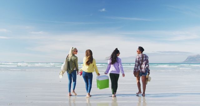 Four friends walking along the shoreline on a sunny day, carrying a cooler and beach gear. Useful for themes of friendship, vacation, summer activities, group outings, and beach holidays.