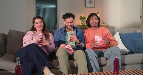 Three friends enjoying cozy late-night movie on sofa with popcorn cups and bottled drinks