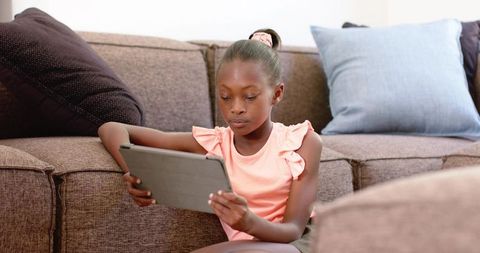 Preteen african girl using tablet on sectional sofa, focused at-home learning and leisure